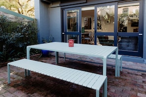 Een witte tafel met een bankje buiten, met een rode emmer op de tafel.