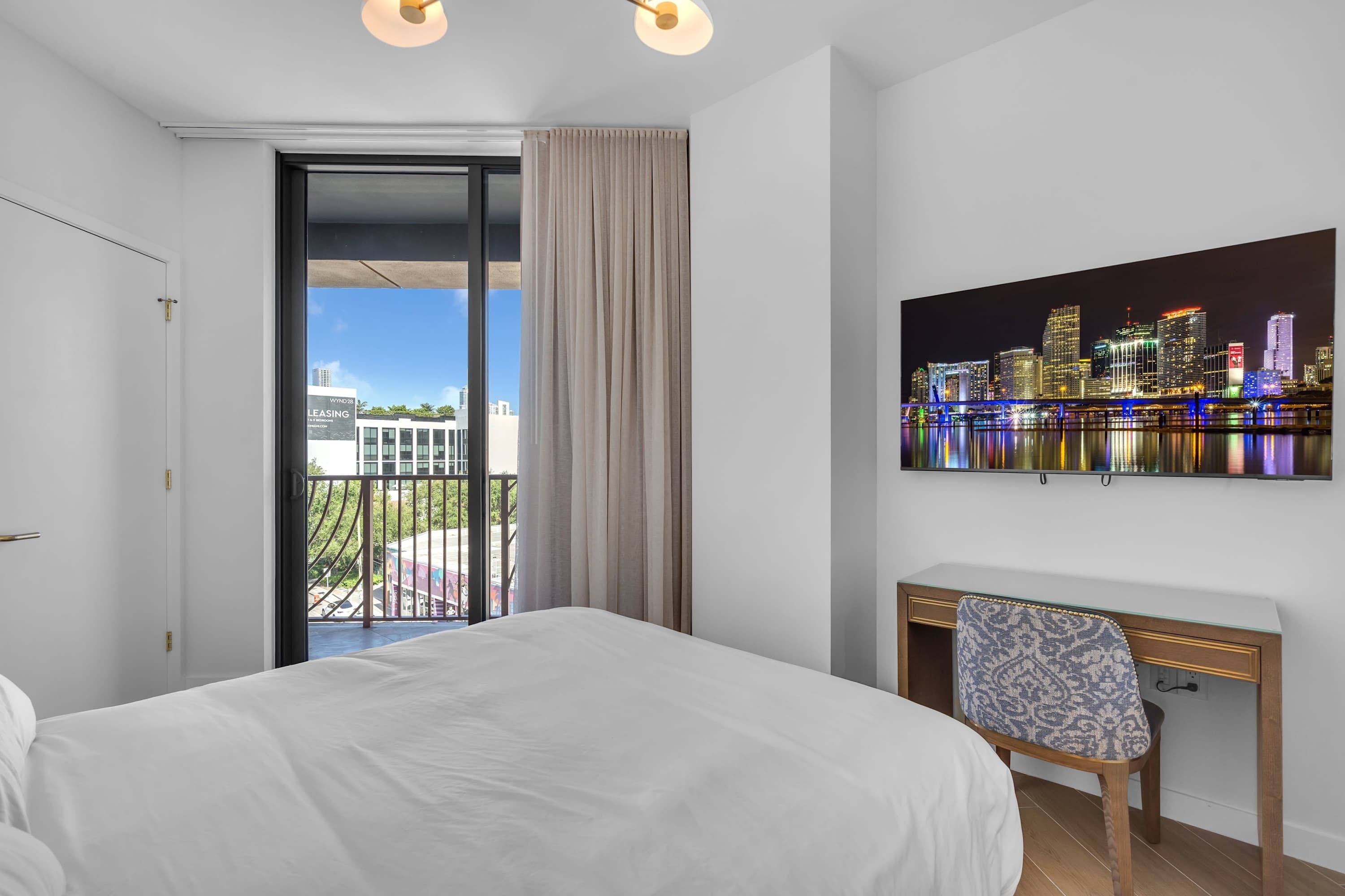 Hotelkamer met een bed, een bureau, een stoel, een stadslandschap schilderij en uitzicht op de stad vanaf een balkon.