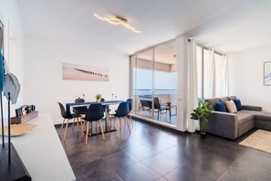 A modern living room with a grey sofa, a dining area with a table and chairs, and a large sliding glass door leading to a balcony. - GuestReady - Oceanview Retreat in Caniço (Caniço de Baixo)
