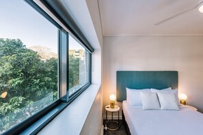 Ein Hotelzimmer mit Bett, Nachttischlampen, einem Fenster mit Blick auf Bäume und einem Deckenventilator.