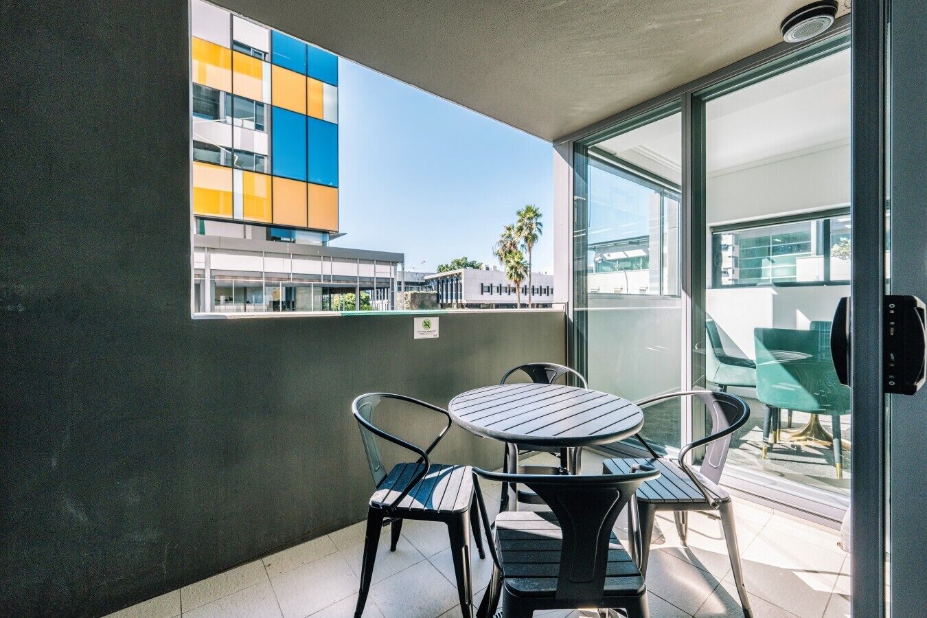 A balcony with a table and chairs, a view of a modern building, and a door to the right.