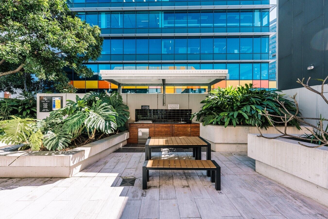 Un espace patio extérieur avec une table en bois et des bancs, entouré de jardinières contenant des plantes tropicales, devant un bâtiment moderne doté de grandes fenêtres en verre.