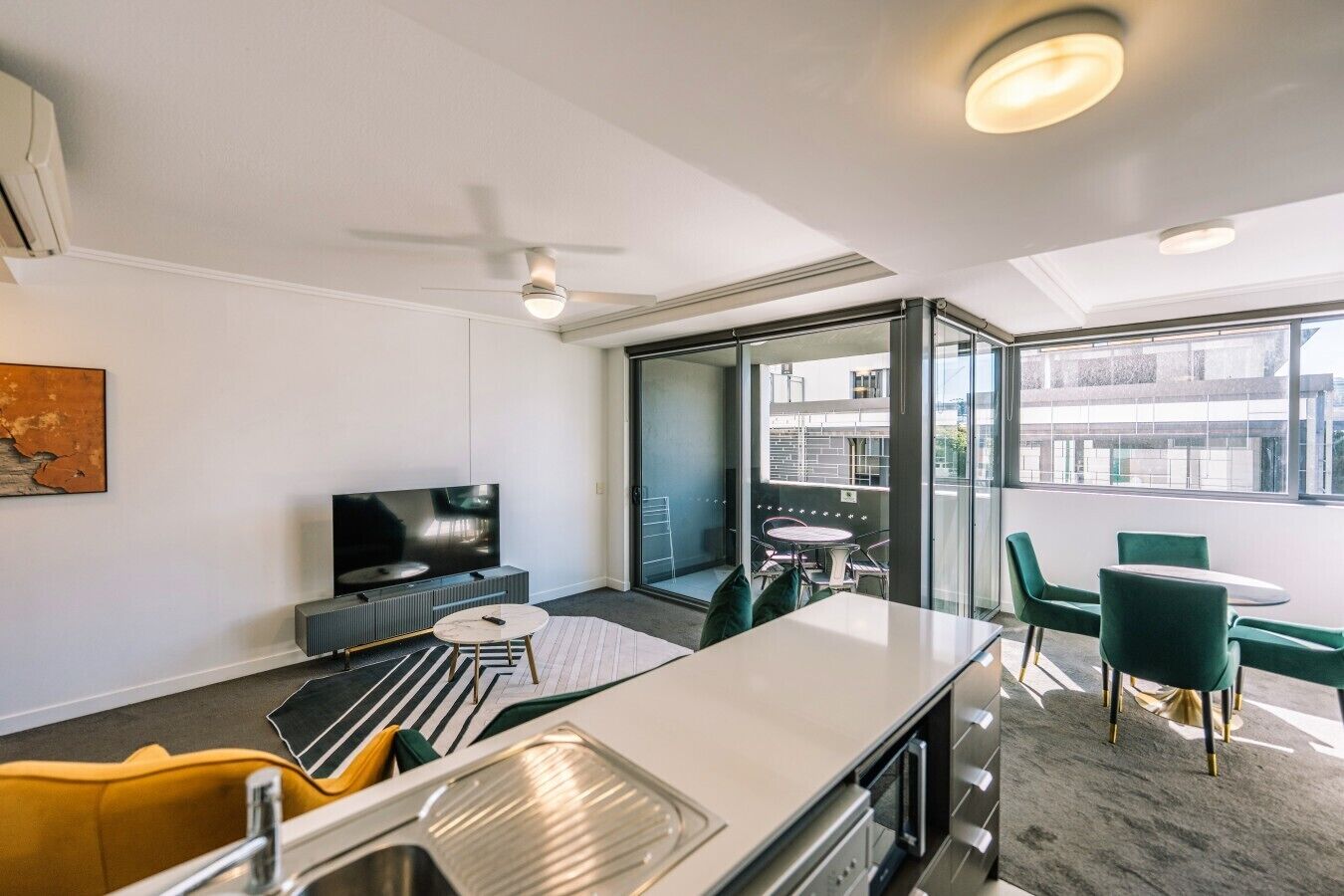 A modern hotel room with a flat-screen TV, a dining area with a round table and chairs, a kitchenette with a sink, and a balcony with a glass door.