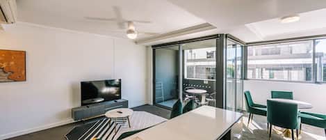 A modern hotel room with a flat-screen TV, a dining area with a round table and chairs, a kitchenette with a sink, and a balcony with a glass door.