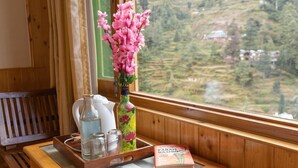 A wooden bench, a tray with a book, a vase with pink flowers, and a window with a view of terraced fields.