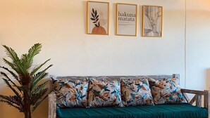 A wooden sofa with floral cushions, a woven rug, and a potted plant in a room with white walls and ceiling. - Samara Studio Beachfront Apartment - Diani Beach (Diani Beach)