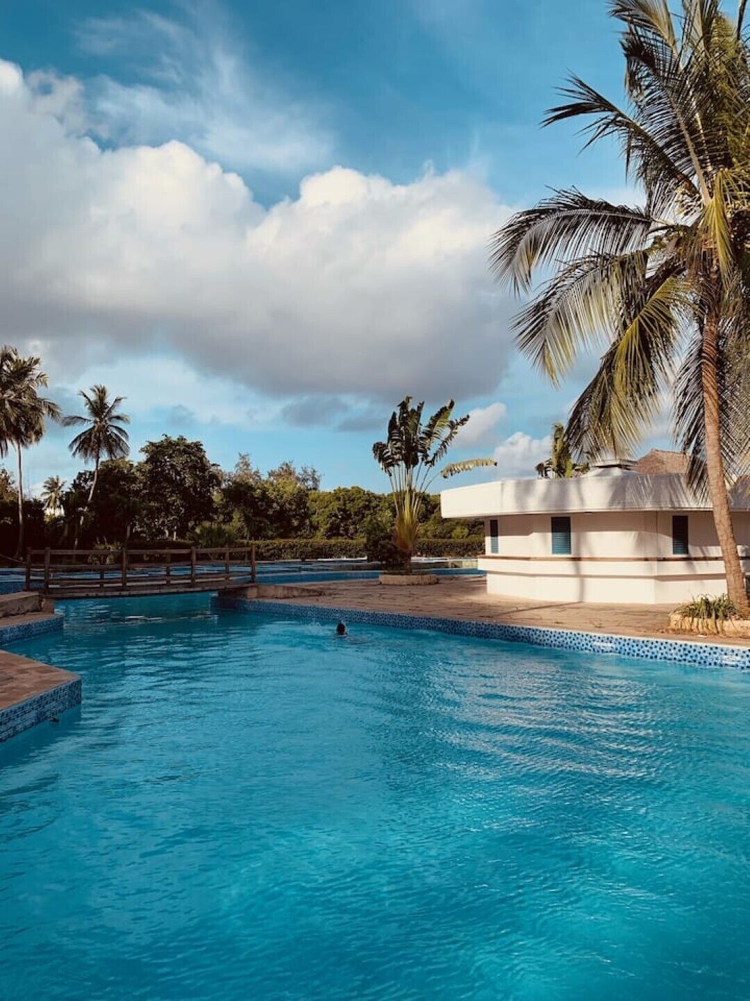 A swimming pool with a wooden bridge, surrounded by palm trees and a building with a white facade.