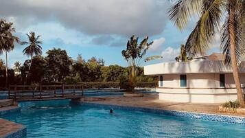 A swimming pool with a wooden bridge, surrounded by palm trees and a building with a white facade.