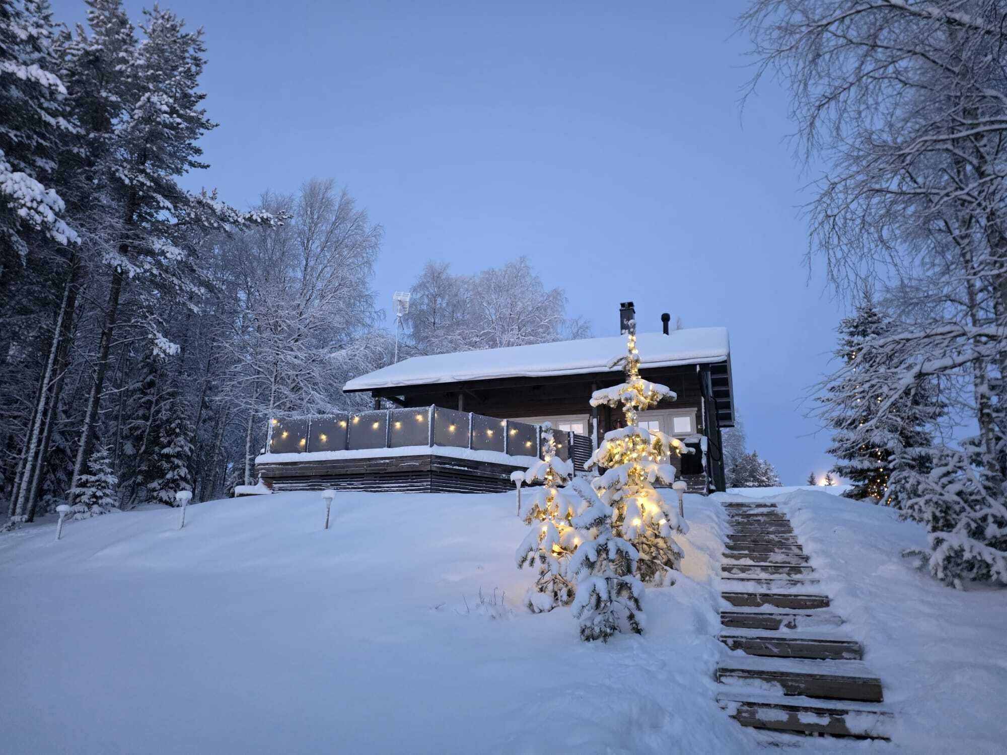 Lumipeitteinen maisema, jossa on näkyvissä puinen mökki ja valaistu joulukuusi.