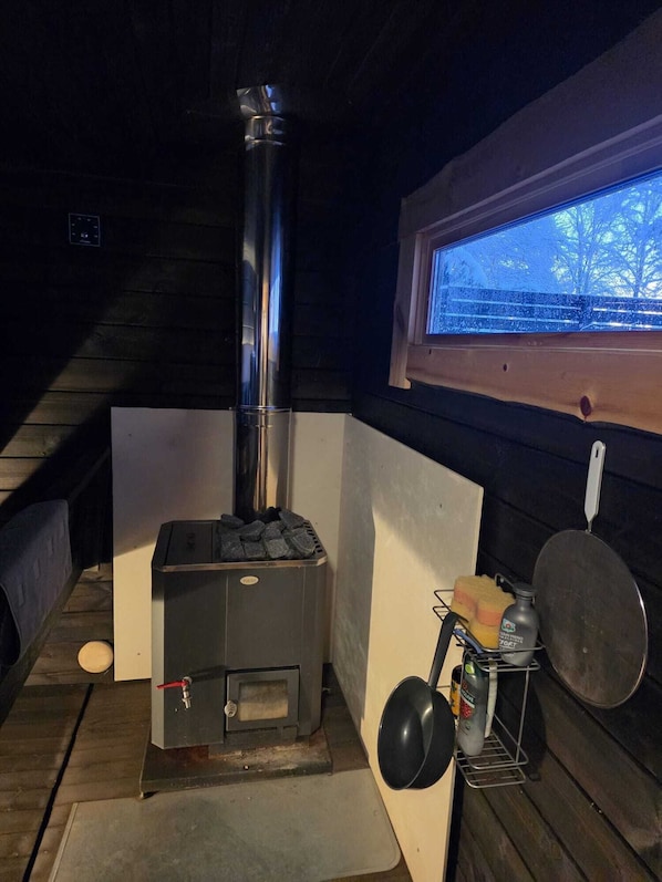 Una estufa de leña con chimenea, una ventana con vistas y una repisa metálica con una olla y una sartén.