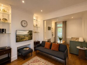 A living room with a sofa, a coffee table, a bookshelf, a clock, and a painting. - Large family house 5 mins walk from station free parking (London)
