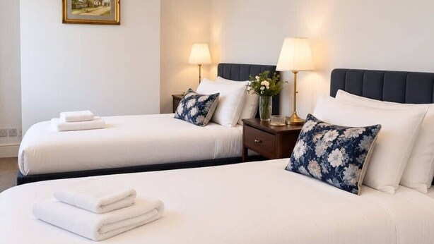 Ein Hotelzimmer mit zwei Betten, jeweils mit weißer Bettwäsche und Kissen, einem Nachttisch mit Lampe sowie einem gerahmten Bild an der Wand.