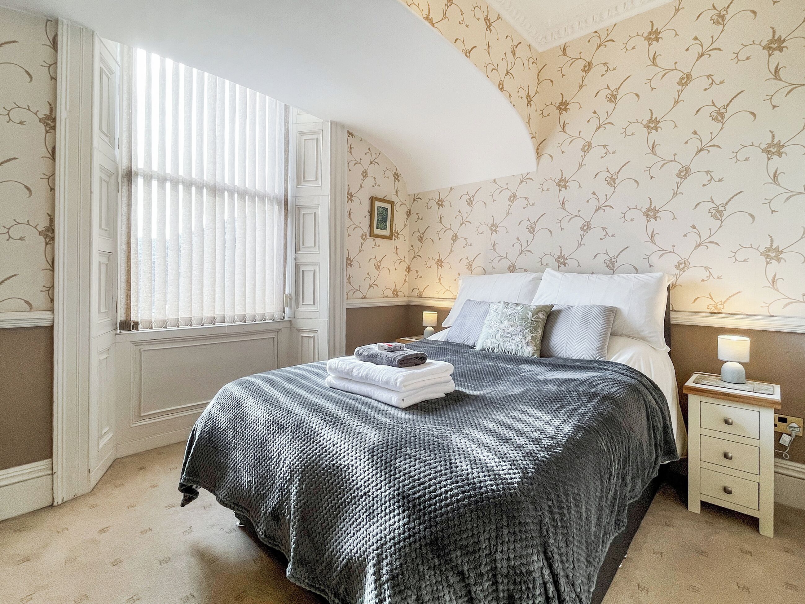 A bedroom with a large bed, a bedside table with a lamp, and a window with blinds.