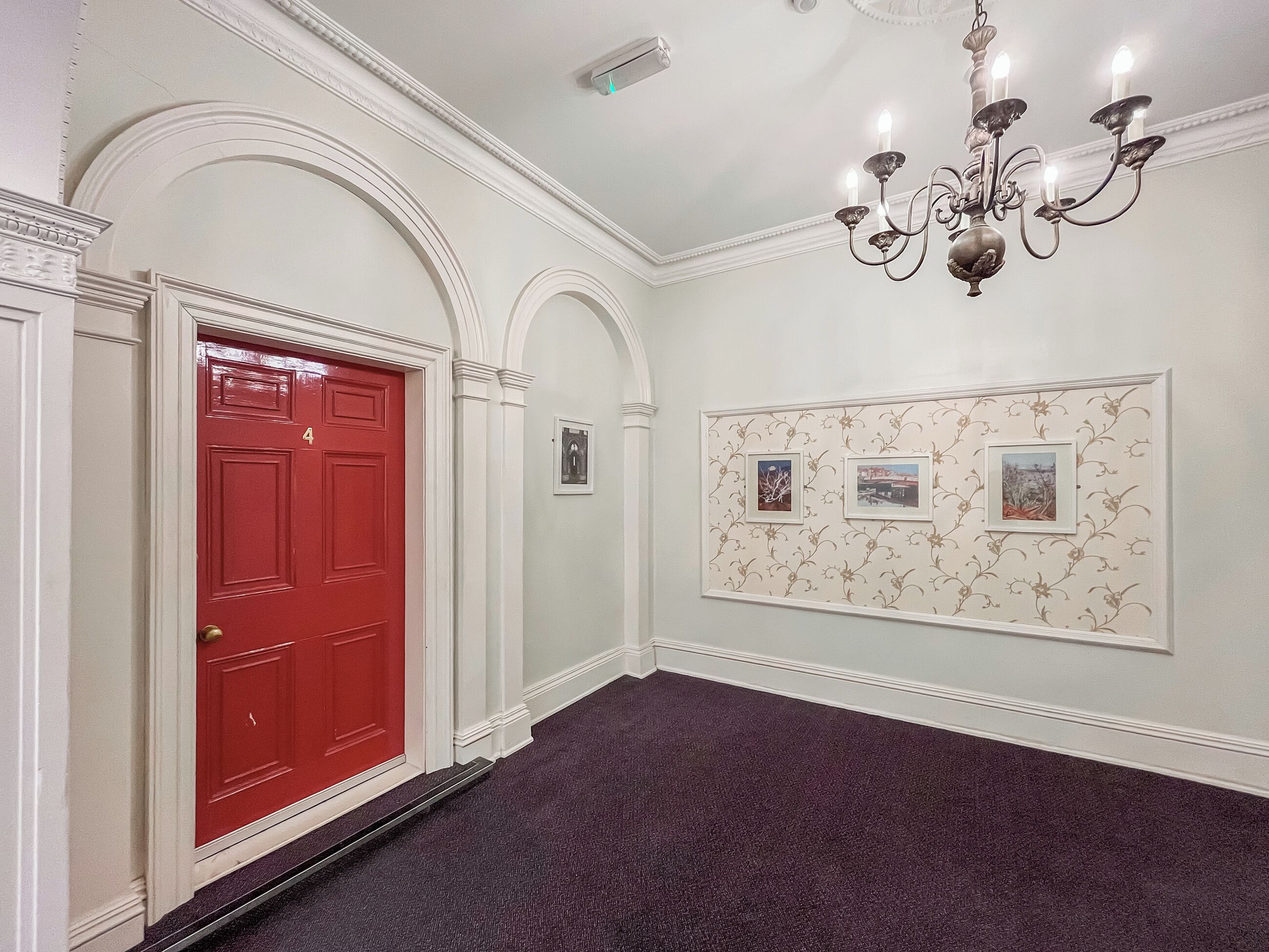 A hallway with a red door, white walls, and a chandelier.