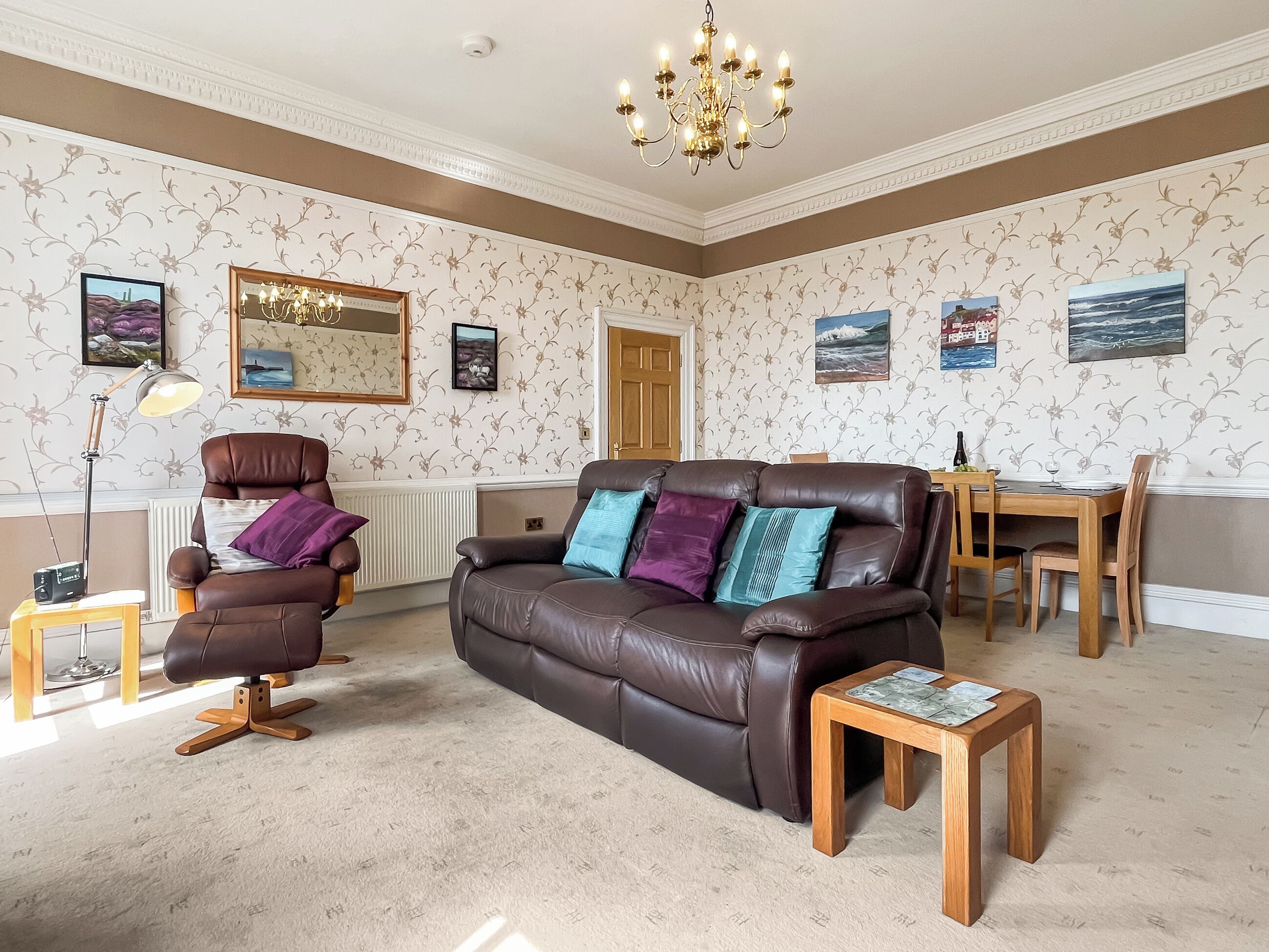 A living room with a brown leather sofa, a wooden coffee table, a dining table with chairs, a standing lamp, and framed artwork on the walls.