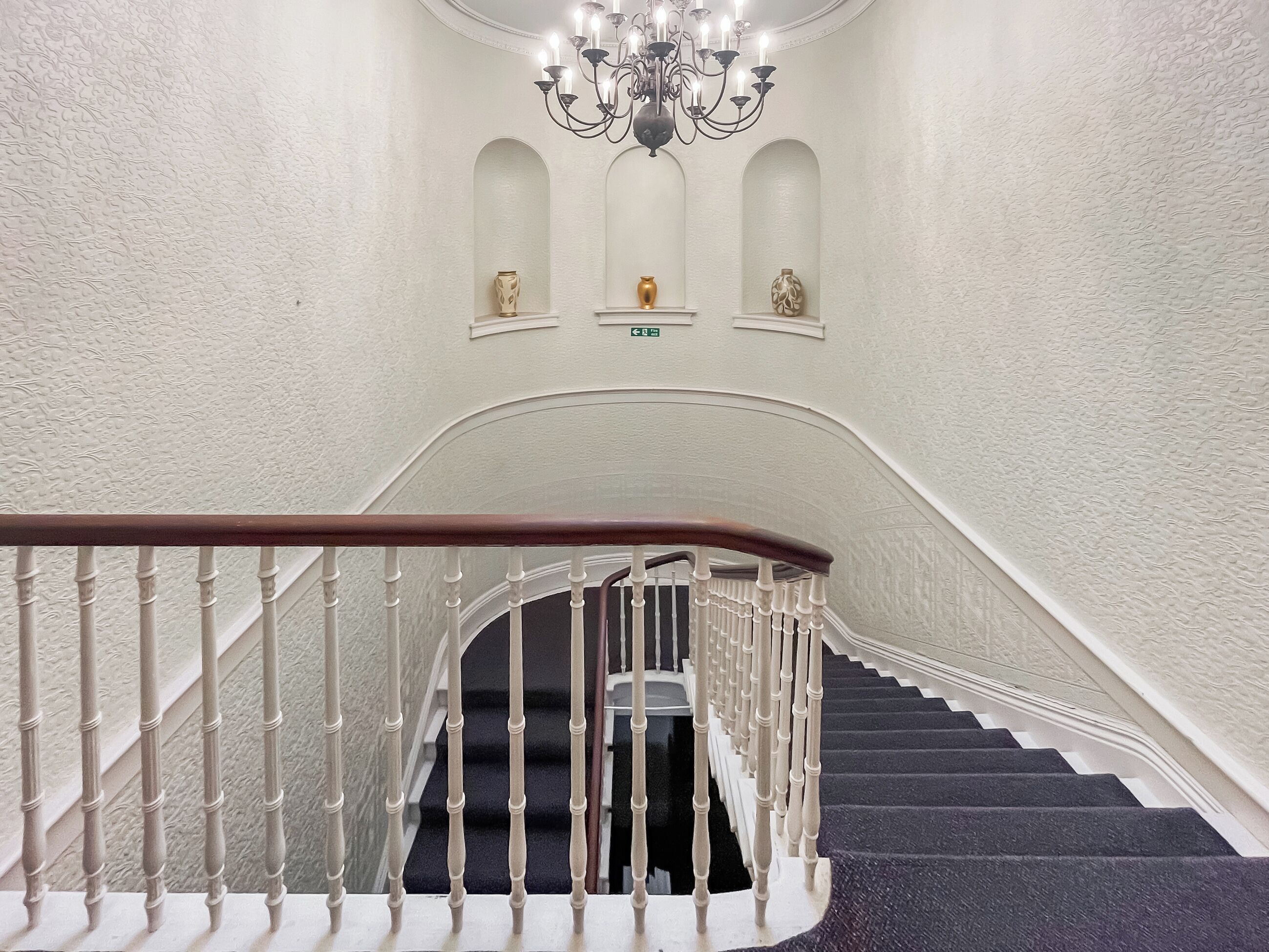 A grand staircase with white balusters, a dark carpeted landing, and a chandelier.