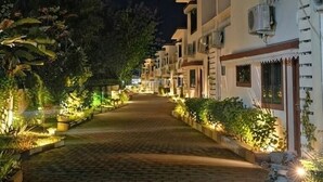Uma trilha pavimentada conduz a uma fileira de edifícios com aparelhos de ar condicionado, em meio a vegetação bem iluminada.