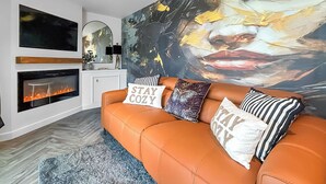 Een woonkamer met een open haard, een groot abstract schilderij en een bank met decoratieve kussens.