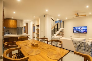 Een moderne woonruimte met een houten eettafel, stoelen, een bank, een aan de muur bevestigde televisie en een trap.