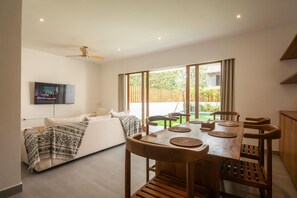 Uma sala de estar moderna com um sofá, uma mesa de jantar com cadeiras, uma TV fixada na parede e uma grande janela com vista para o exterior.