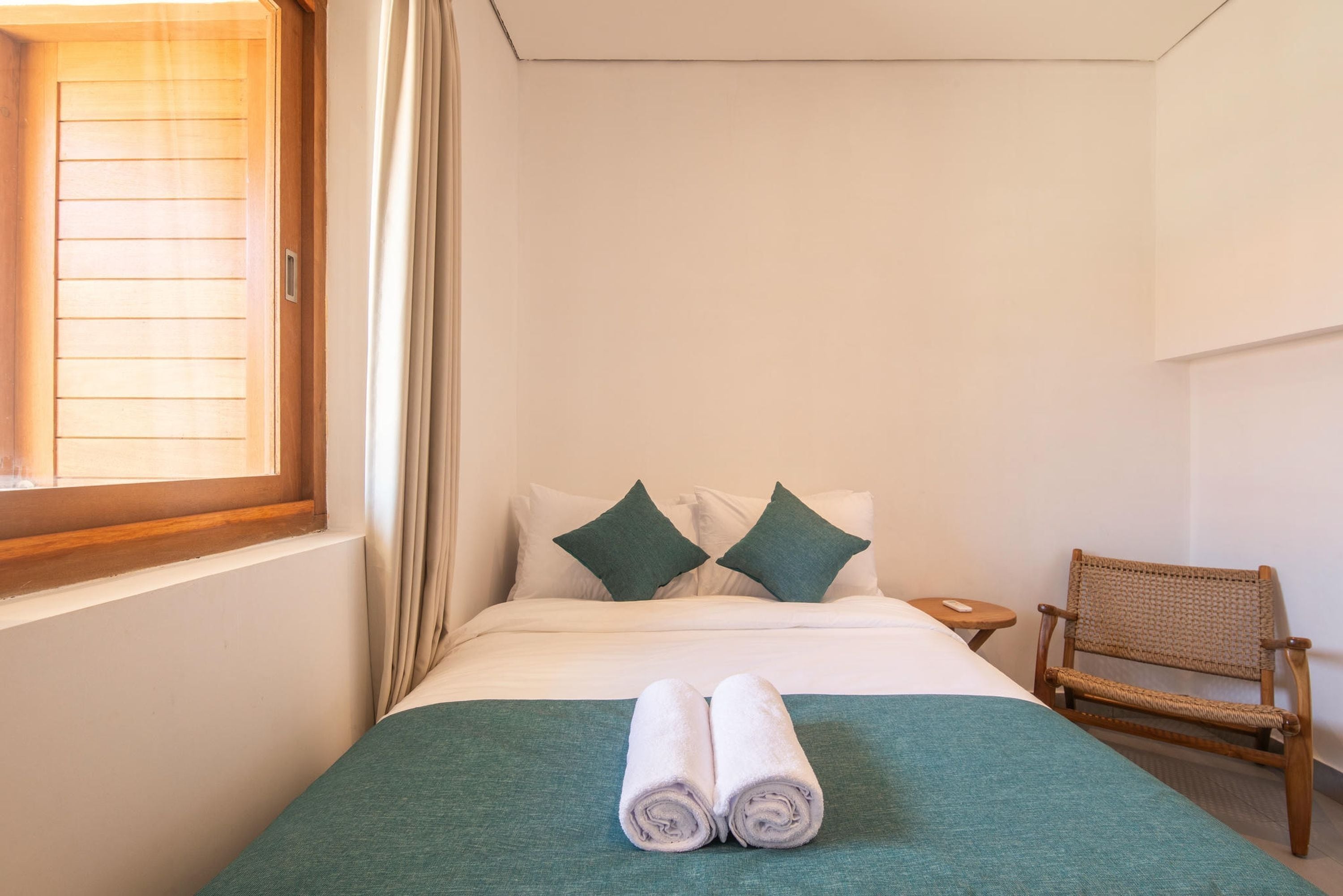 Quarto de hotel com cama, duas almofadas, mesa de cabeceira pequena, uma cadeira e janela com cortinas.