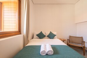 Quarto de hotel com cama, duas almofadas, mesa de cabeceira pequena, uma cadeira e janela com cortinas.