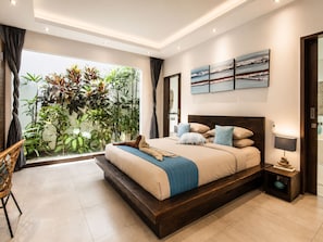 A modern bedroom with a large bed, a view of greenery through a window, and a nightstand with a lamp.