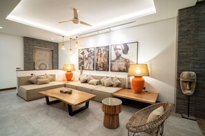 Ein modernes Wohnzimmer mit einer Sofagarnitur, einem Couchtisch und einem Sessel. An den Wänden sind Lampen angebracht und es gibt einen Deckenventilator.