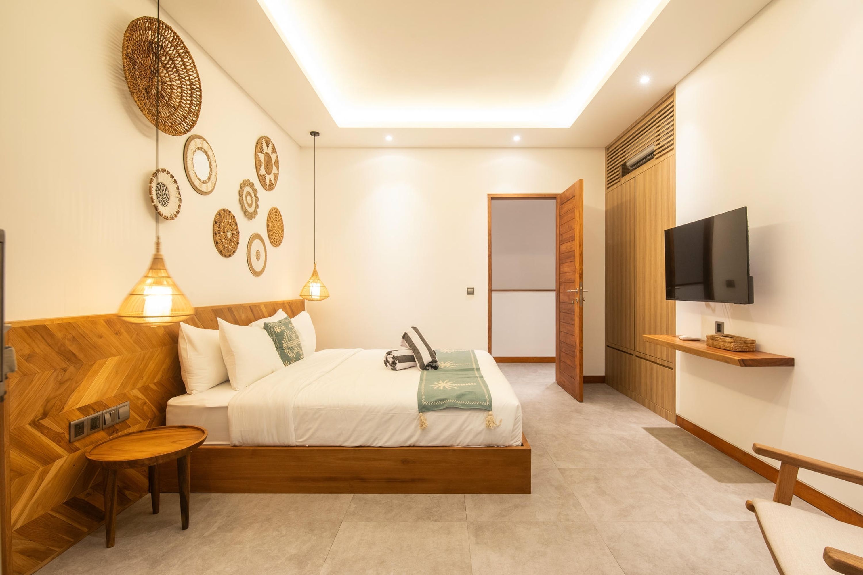 Une chambre à coucher moderne avec une tête de lit en bois, un lit avec une literie blanche, une table de chevet, une lampe, un téléviseur fixé au mur et une armoire.