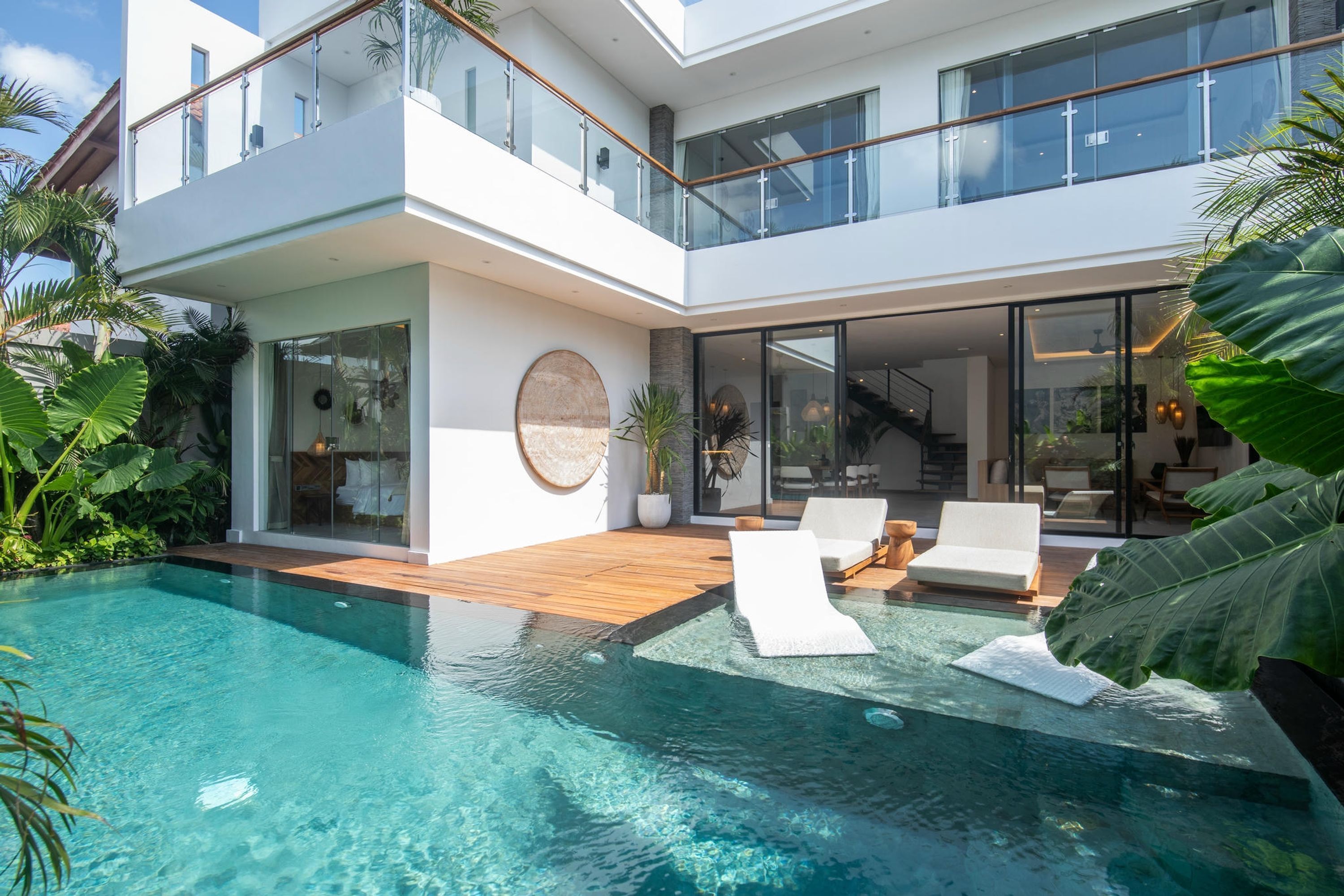 Een modern huis met twee verdiepingen, een glazen balkon, een zwembad en een terras met lounge stoelen.