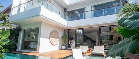 Una casa moderna de dos pisos con balcón de cristal, piscina y plantas tropicales.