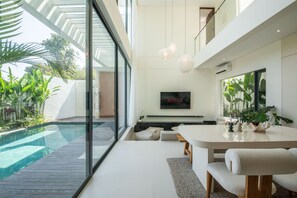 A modern living room with a large TV, a dining area, and a glass wall overlooking a swimming pool. - Casa Rio-beautiful 3br Villa Center Seminyak (Keroboken Kelod)