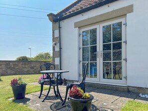 A patio with a table and chairs, a potted plant, and a building with a large window and a door. - 3 bedroom accommodation in Beadnell (Beadnell)