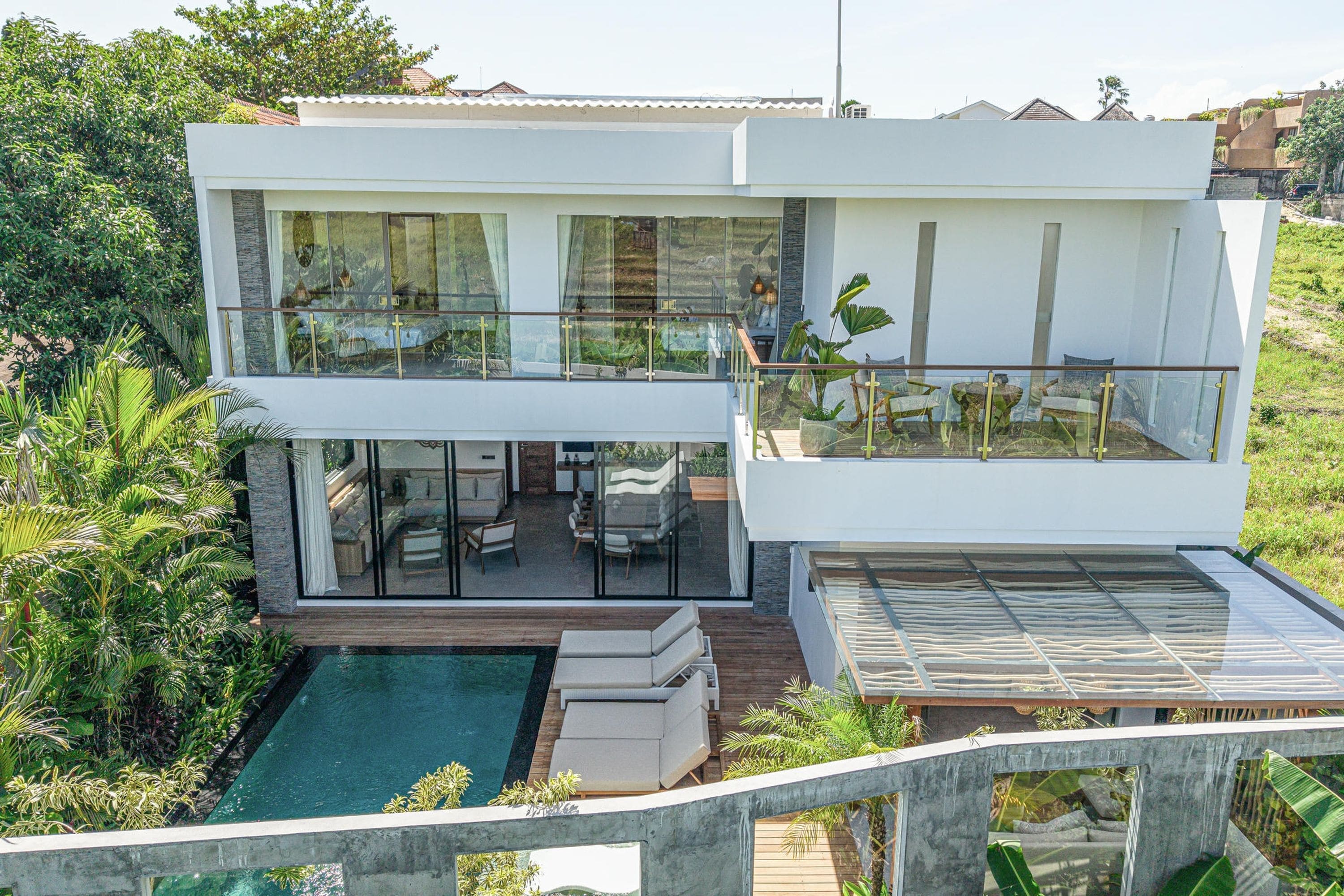 A modern two-story house with a glass balcony, a pool, and lush greenery.