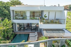 Een modern huis met twee verdiepingen, een glazen balkon, een zwembad en weelderige begroeiing.