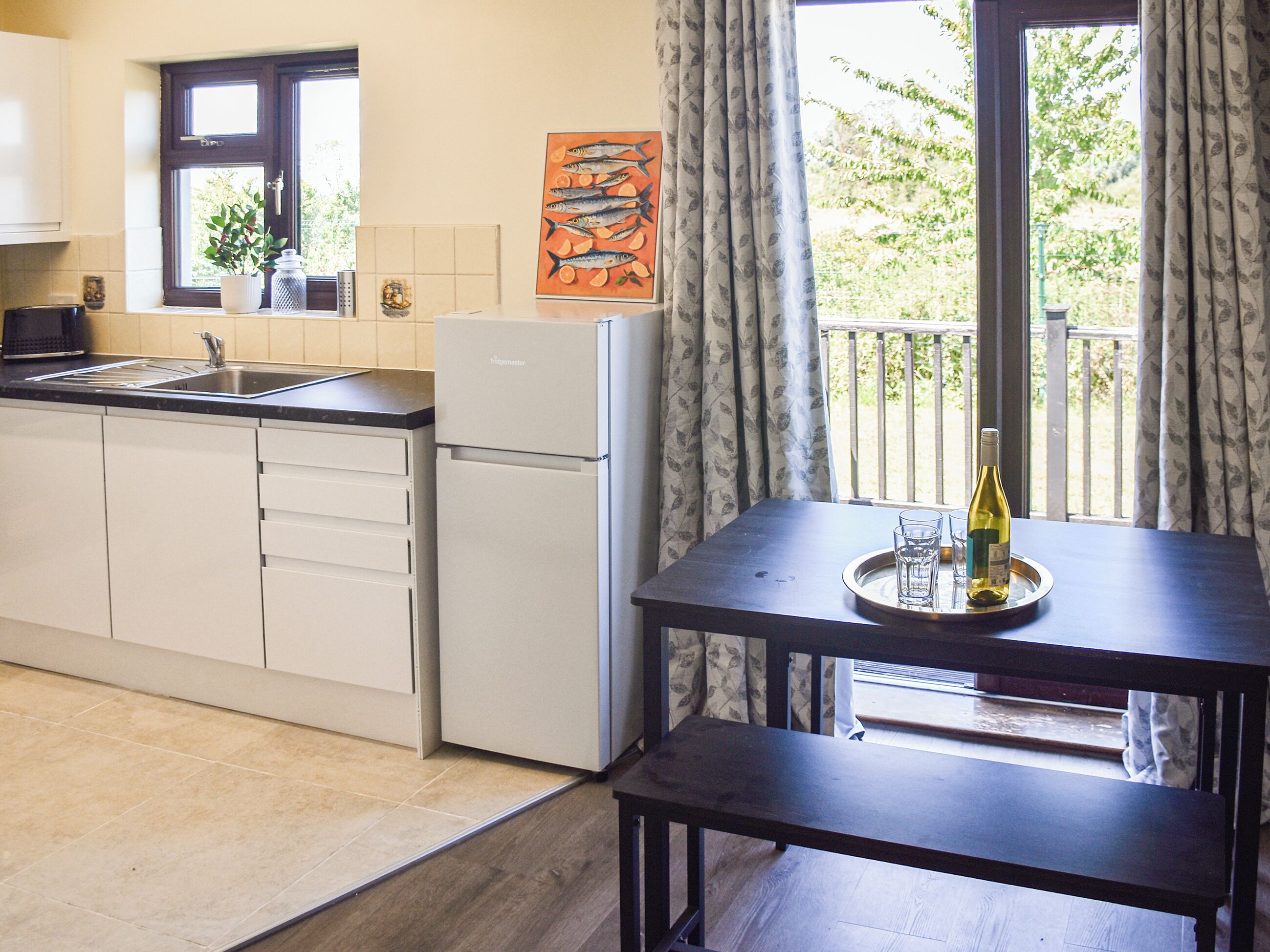 A kitchenette with a refrigerator, sink, and cabinets, a dining table with a tray holding glasses and a bottle, and a window with curtains.