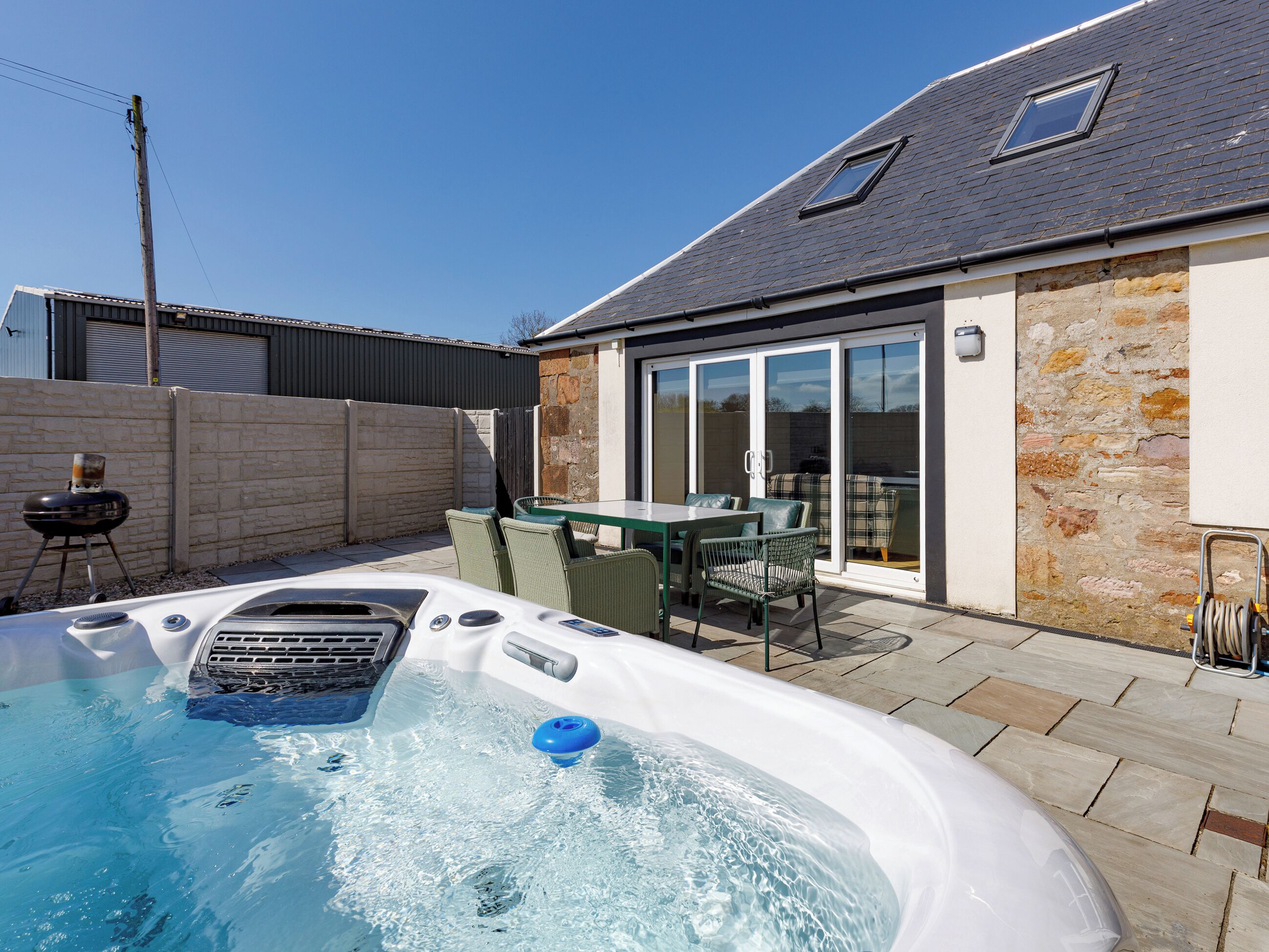 Un jacuzzi dans un jardin, avec une table et des chaises, un barbecue et un mur en pierre.