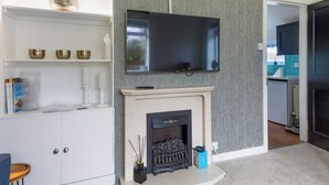 A modern living room with a fireplace, a flat-screen TV mounted on the wall, and a white entertainment unit with decorative items. - The Paddock - UK52288 (Auchincruive)