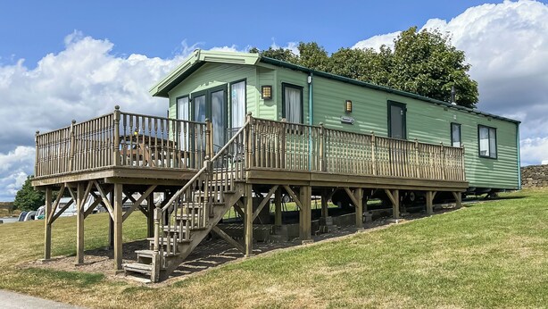 Una casa móvil verde con terraza de madera y escaleras, ubicada en una colina cubierta de césped.