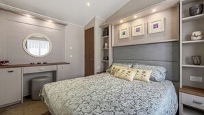 Un dormitorio con una cama, un tocador con espejo y estanterías empotradas con objetos decorativos.