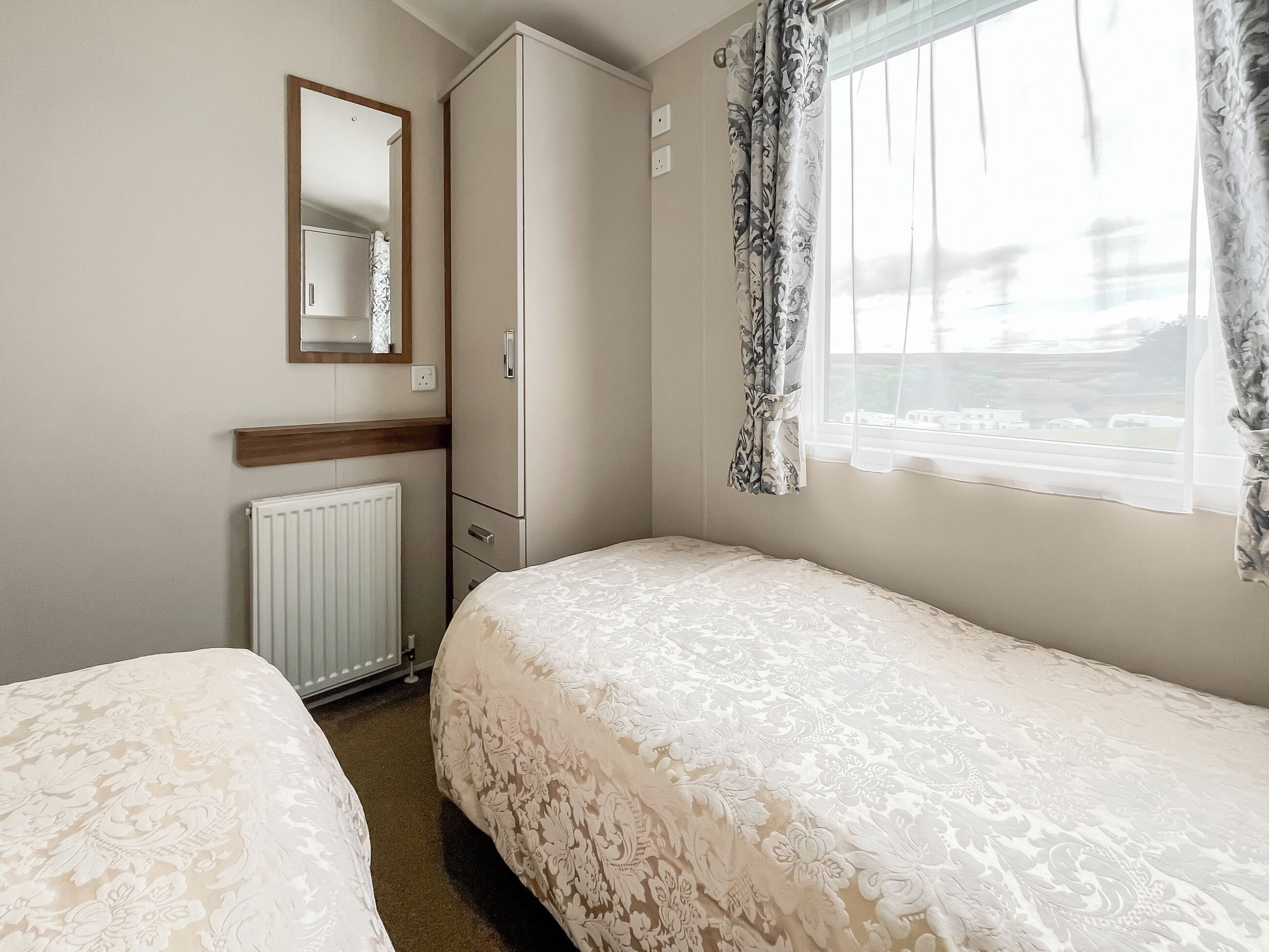 A bedroom with two beds, a wardrobe, a mirror, and a window with curtains.