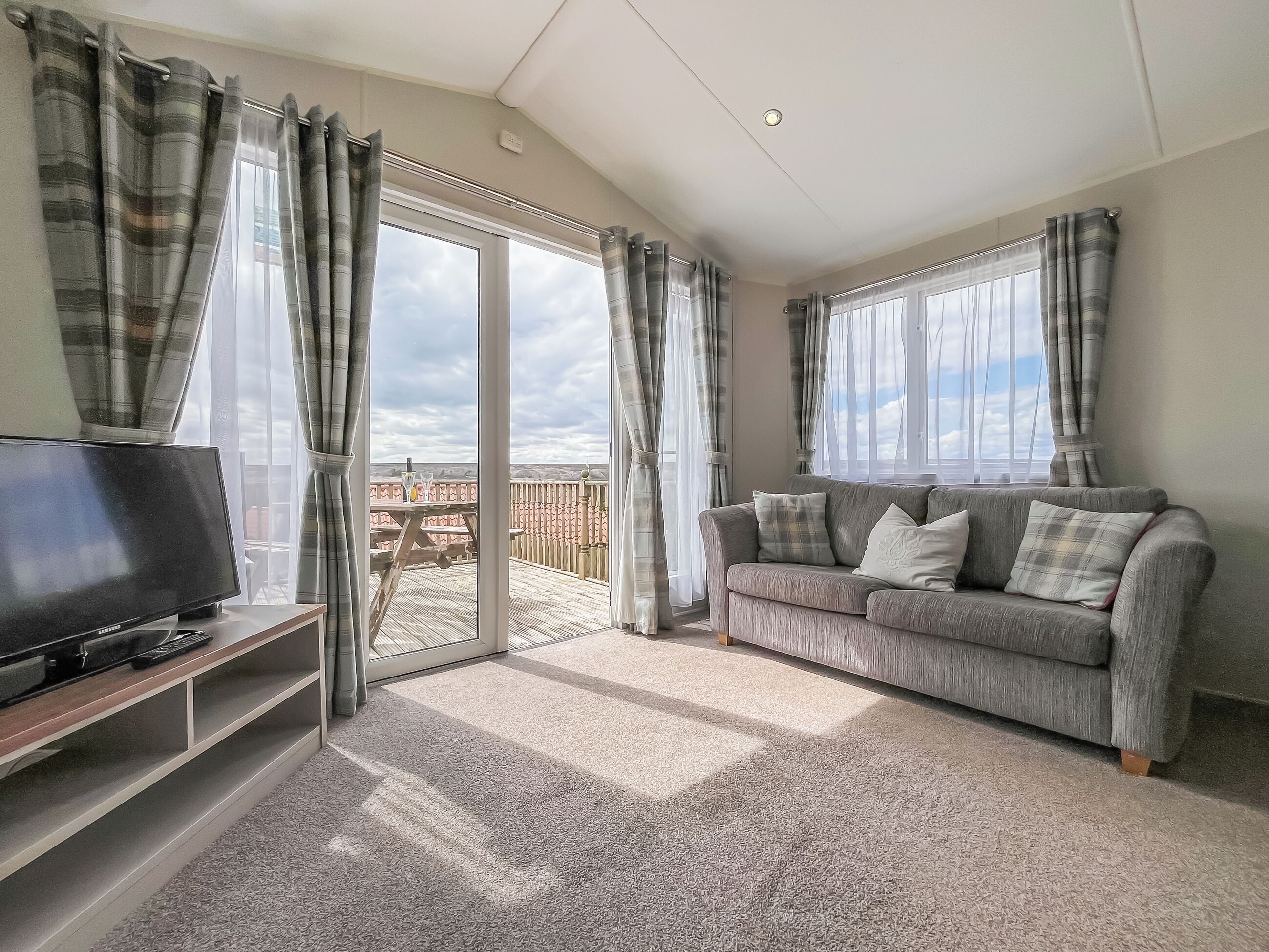 A living room with a grey sofa, a flat-screen TV, and a balcony with a wooden deck.