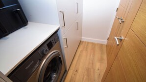 A compact laundry room with a front-loading washing machine, wooden cabinets, and a white countertop. - 6 bedroom accommodation in Carnforth (Carnforth)