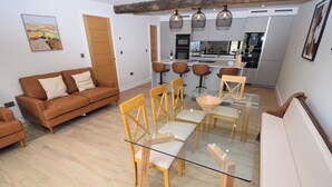 A modern living room with a glass dining table, wooden chairs, a sofa, and a kitchen in the background. - 6 bedroom accommodation in Carnforth (Carnforth)