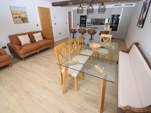 Uma sala moderna com uma mesa de jantar de vidro, cadeiras de madeira, um sofá e uma cozinha ao fundo.