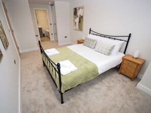 Uma cama bem arrumada com um edredom verde e lençóis brancos, uma mesinha de cabeceira de madeira com uma luminária e um quadro na parede.