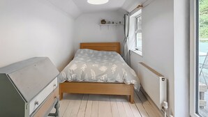 Una habitació estreta amb un llit, una petita còmoda, un radiador i una finestra.