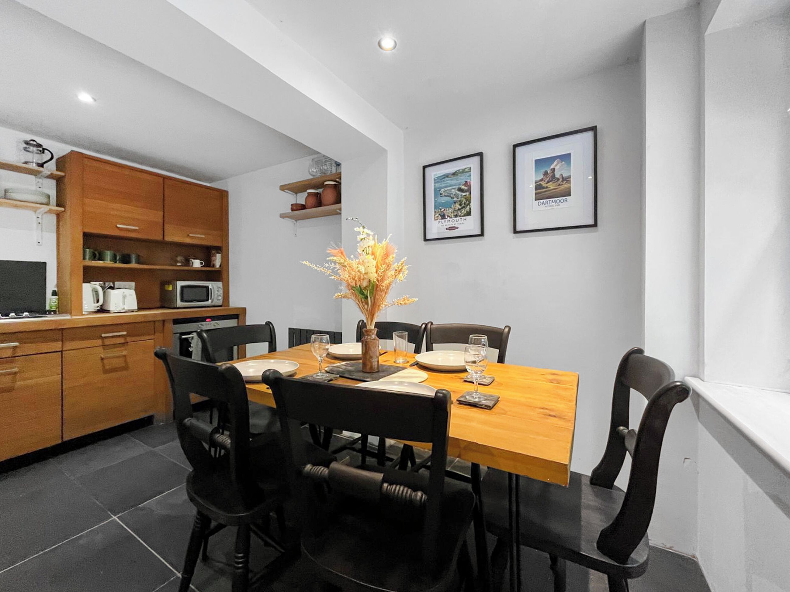 Comedor con una mesa de madera, sillas negras y un jarrón de flores. Se observan cuadros en la pared y una cocina con armarios de madera y electrodomésticos.