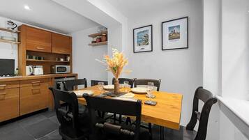 Comedor con una mesa de madera, sillas negras y un jarrón de flores. Se observan cuadros en la pared y una cocina con armarios de madera y electrodomésticos.