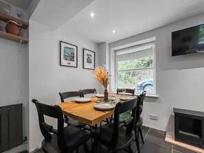 A dining area with a wooden table, black chairs, and a vase with dried flowers. There are framed pictures on the wall and a window with a view of greenery and a car outside.